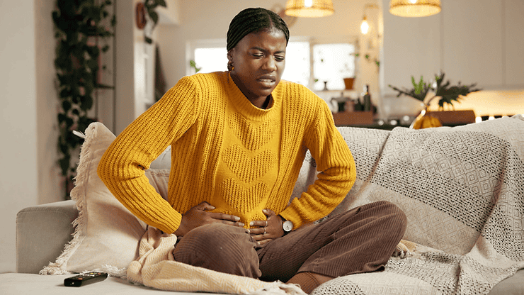 Woman in yellow sweater sitting on a couch, holding her stomach in discomfort, illustrating symptoms of Irritable Bowel Syndrome (IBS) and gut health issues.
