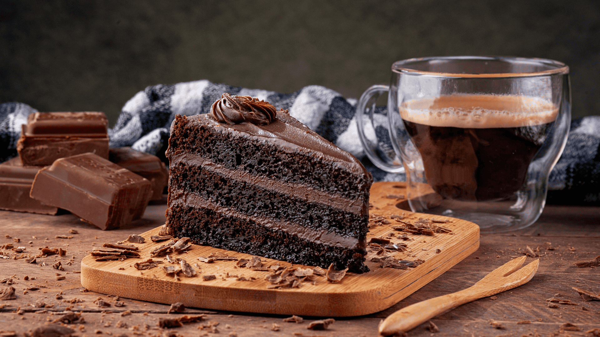 Chocolate cake slice with rich frosting on wooden board, surrounded by chocolate bars, chocolate shavings, and a cup of coffee, illustrating indulgent dessert options.