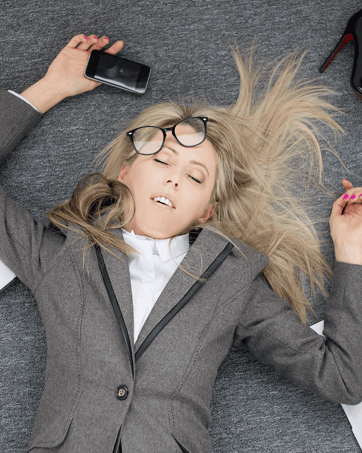 Woman in business attire lying on the floor, holding a smartphone, with glasses on her face, representing stress and burnout related to IBS.