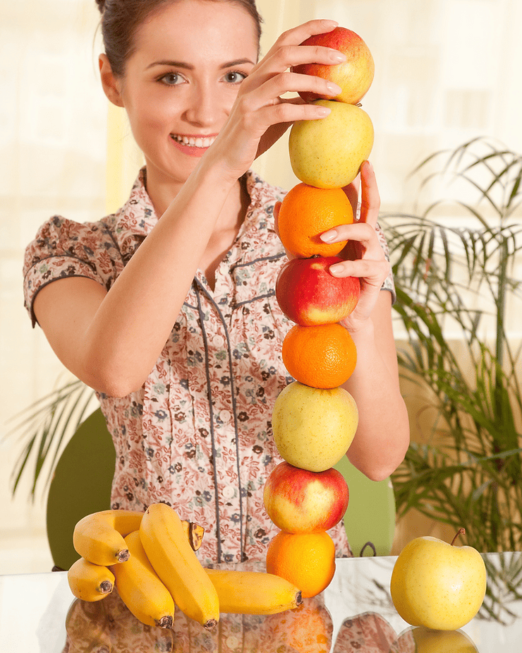 Woman stacking colorful fruits (apples, oranges, and a pear) while bananas are arranged beside her, illustrating FODMAP stacking concepts in relation to gut health.