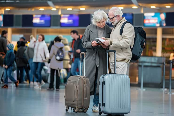 Elderly couple at airport reviewing travel plans, surrounded by travelers and luggage, emphasizing travel challenges for individuals with IBS.