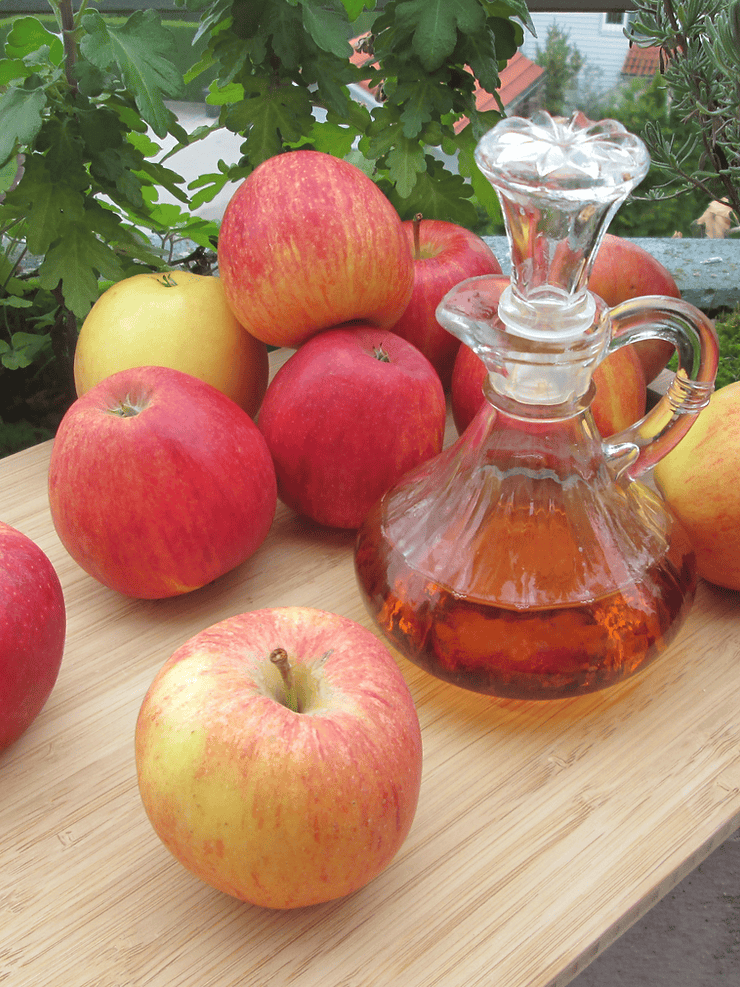 Assorted apples and a glass decanter of vinegar on a wooden surface, symbolizing the health benefits of vinegar for digestion and blood sugar regulation.
